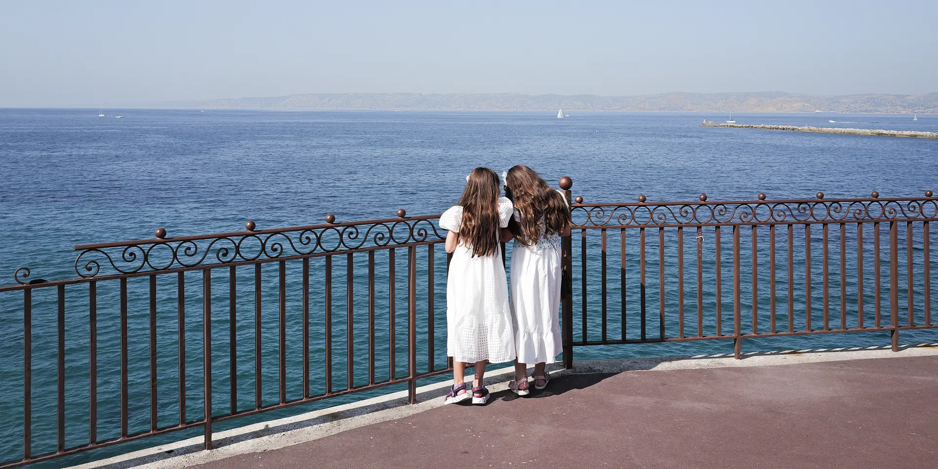 Two girls in white dresses stand at a railing and look to the sea.